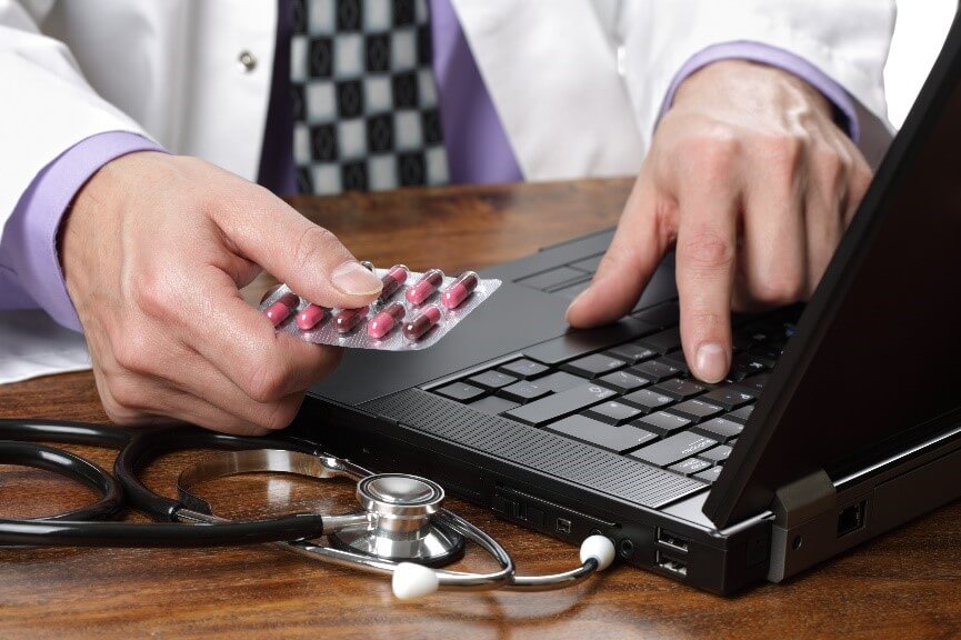 Stock photo of doctor holding pills in one hand while typing on a laptop with the other